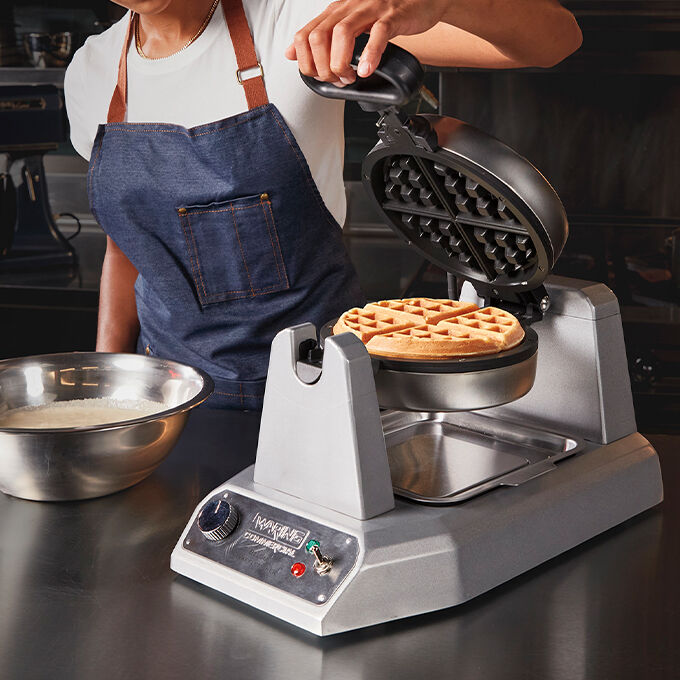 Waring Belgian Waffle maker being opened in a commercial kitchen setting with a cooked waffle inside.