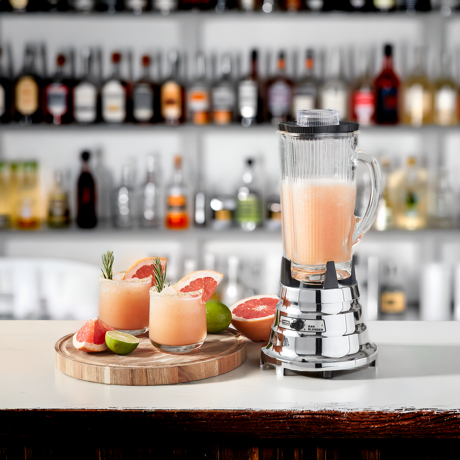 silver blender sitting on a bar with 2 pink blended drinks and fruit next to it.